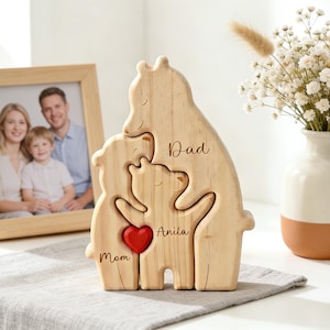 Op de afbeelding: Houten familieberenpuzzel met de woorden "Papa", "Mama" en "Anita" gegraveerd. Een rood hart staat in het midden. Een ingelijste familiefoto en een vaas met bloemen staan op de achtergrond. De houten berenpuzzel is een decoratief item.