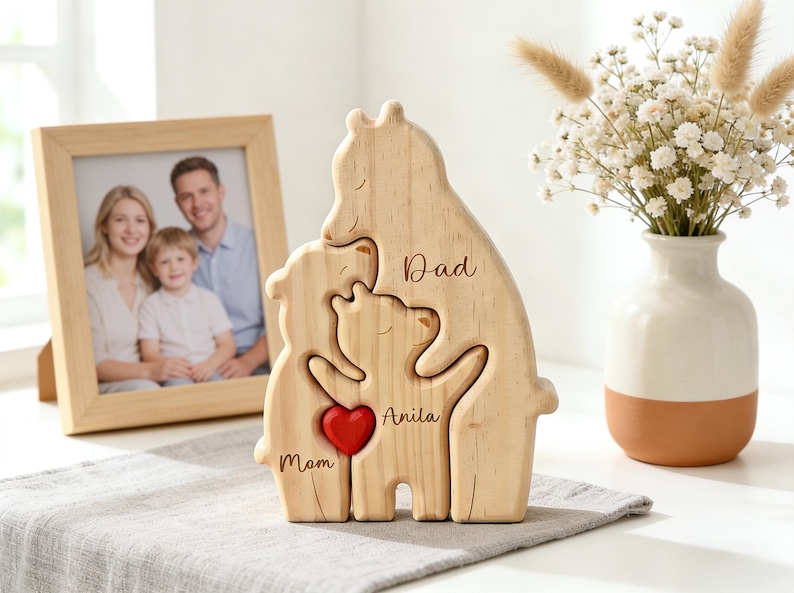 Op de afbeelding: Houten familieberenpuzzel met de woorden "Papa", "Mama" en "Anita" gegraveerd. Een rood hart staat in het midden. Een ingelijste familiefoto en een vaas met bloemen staan op de achtergrond. De houten berenpuzzel is een decoratief item.