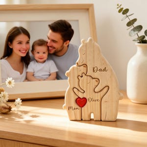 Op de afbeelding: Houten berenfamiliebeeldje met de woorden "Papa", "Mama" en "Olive" en een rood hart. Een ingelijste familiefoto staat op de achtergrond. Een vaas met groen en bloemen is ook zichtbaar.