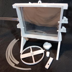 May include: A white, rectangular, transparent-windowed structure with a clear tube extending from the side. Additional components include curved tubes, a circular frame, a small cylinder, and a rectangular piece, all in white.