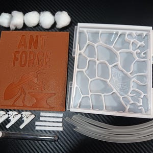 May include: An ant farm kit with various components. Includes a brown rectangular piece with "ANT FORGE" and an ant graphic, a clear plastic ant habitat, cotton balls, a pipette, tubes, and other accessories.