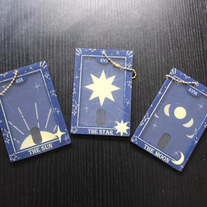 Op de afbeelding: Drie blauwe tarotkaartsleutelhangers met gouden kettingbevestigingen. Elke kaart heeft een ander ontwerp: De Zon, De Ster en De Maan. De kaarten hebben witte illustraties en tekst, met een ster- en maanontwerp.