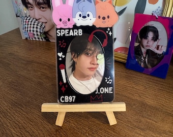 May include: A black photo card featuring a person, with the text "J.ONE" and "CB97". The card is displayed on a small wooden easel. Above the card are three cartoon animal charms: a pink pig, a blue dog, and an orange fox. A magazine and two framed photos are in the background.