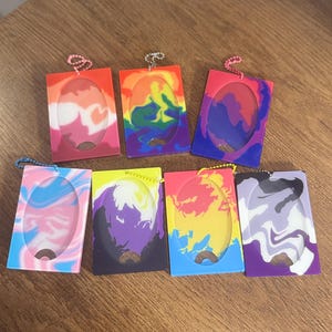 May include: A collection of seven rectangular silicone cardholders with oval cutouts. Each holder features a unique swirled color pattern, including rainbow, pink, blue, purple, yellow, and black. They have a small metal chain and a loop for attachment.
