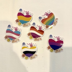 May include: A collection of heart-shaped stickers featuring pride flag designs. Each sticker has a different flag pattern, including rainbow, transgender, lesbian, non-binary, and bisexual flags, with cute bunny and bear illustrations.