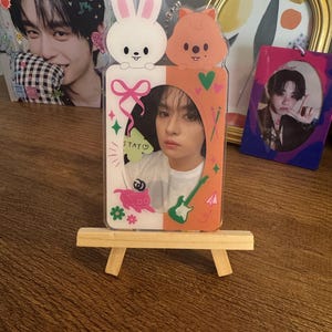 May include: A clear acrylic photo frame with a cartoon bunny and squirrel design at the top. The frame holds a photo of a person. The frame is on a small wooden easel. Other photos and a framed picture are in the background.