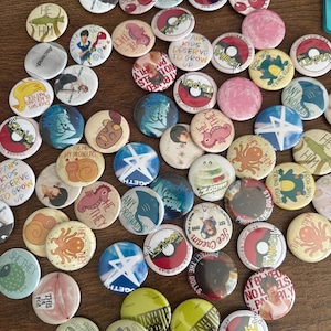May include: A collection of circular pin-back buttons featuring various designs, including illustrations of animals, abstract patterns, and text such as "Trans kids deserve to grow up," "Any pronouns," "The Leader," "The Ace," "The Vocalist," and "Protect the dolls."
