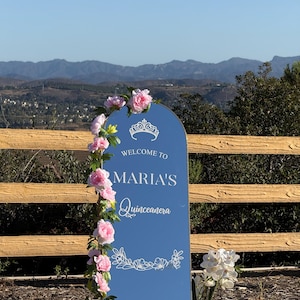 Puede incluir: Un letrero de bienvenida de Quincea&ntilde;era azul con el texto "Welcome to Maria's Quincea&ntilde;era" en escritura blanca. El letrero est&aacute; decorado con flores rosas y una guirnalda floral. El fondo presenta una valla de madera y una cordillera.