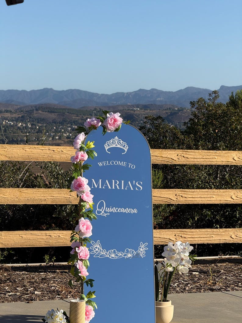 Puede incluir: Un letrero de bienvenida de Quincea&ntilde;era azul con el texto "Welcome to Maria's Quincea&ntilde;era" en escritura blanca. El letrero est&aacute; decorado con flores rosas y una guirnalda floral. El fondo presenta una valla de madera y una cordillera.