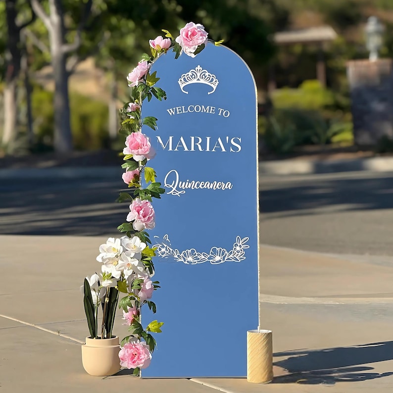 Puede incluir: Un letrero de bienvenida azul para "Maria's Quinceanera" con texto blanco y detalles florales. El letrero est&aacute; decorado con flores rosas y blancas y una peque&ntilde;a planta en maceta. Una vela se encuentra en la base.