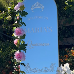Puede incluir: Un letrero espejado con la inscripci&oacute;n "WELCOME TO MARIA'S Quinceanera", adornado con una corona y motivos florales. El letrero est&aacute; enmarcado por flores rosas y blancas y vegetaci&oacute;n, sobre un fondo de follaje.