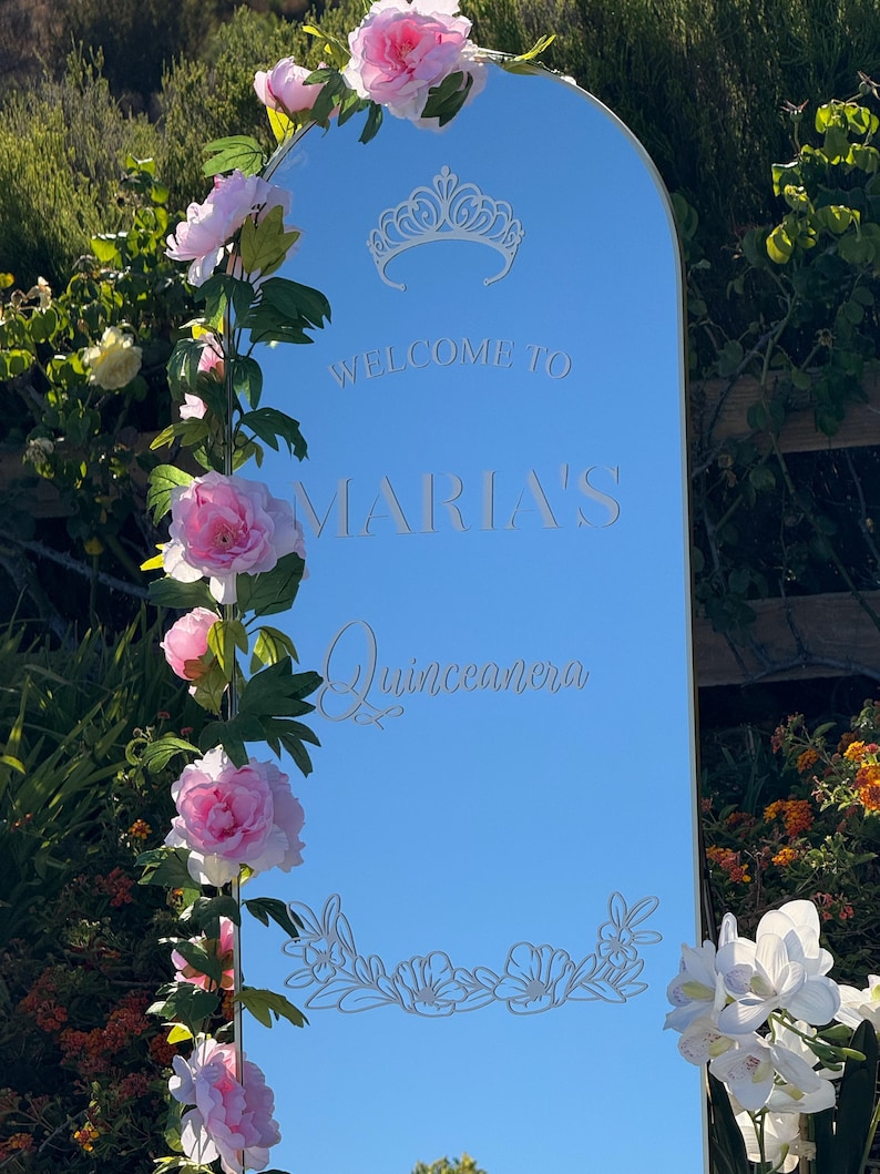 Puede incluir: Un letrero espejado con la inscripci&oacute;n "WELCOME TO MARIA'S Quinceanera", adornado con una corona y motivos florales. El letrero est&aacute; enmarcado por flores rosas y blancas y vegetaci&oacute;n, sobre un fondo de follaje.
