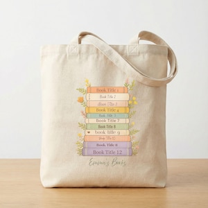 May include: Beige tote bag featuring a stack of colorful books with floral accents. Each book has a title, with the text "Emma's Books" at the bottom. The bag has two handles and is made of a light-colored fabric.