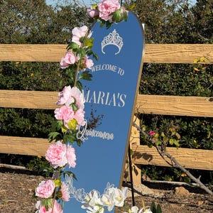 Puede incluir: Un espejo alto y arqueado con marco dorado, adornado con flores rosas y orqu&iacute;deas blancas. El espejo refleja el cielo y presenta el texto "WELCOME TO MARIA'S Quinceanera" en escritura blanca, con un gr&aacute;fico de corona.