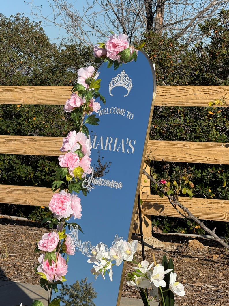 Puede incluir: Un espejo alto y arqueado con marco dorado, adornado con flores rosas y orqu&iacute;deas blancas. El espejo refleja el cielo y presenta el texto "WELCOME TO MARIA'S Quinceanera" en escritura blanca, con un gr&aacute;fico de corona.