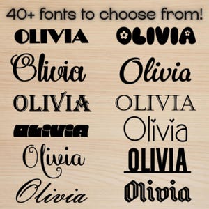 May include: A wooden background displays various black text examples of the name "Olivia" in different fonts. The text "40+ fonts to choose from!" is at the top. The fonts vary in style, from bold to script.