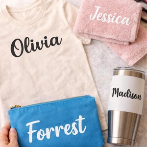 May include: A cream-colored t-shirt with the name Olivia in black, a pink towel with the name Jessica in white, a blue zippered pouch with the name Forrest in white, and a stainless steel tumbler with the name Madison in black text.