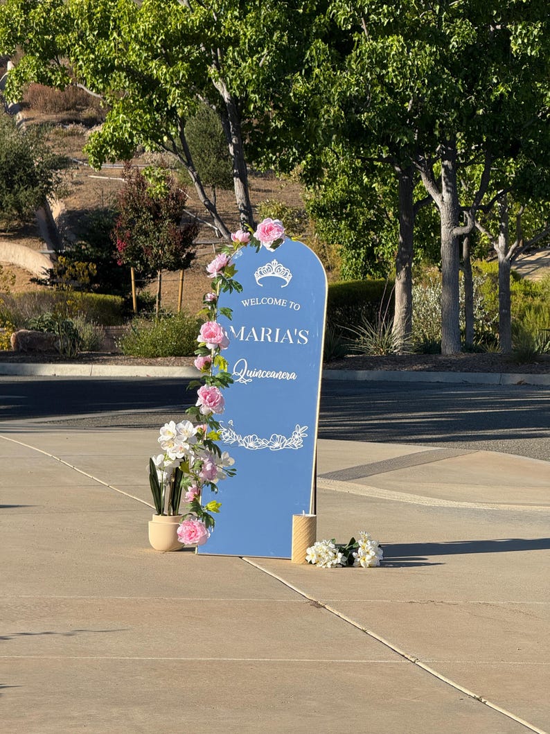 Puede incluir: Un letrero azul con el texto "Welcome to Maria's Quinceanera" est&aacute; decorado con flores rosas y blancas. Un peque&ntilde;o jarr&oacute;n con flores y una vela est&aacute;n junto al letrero. El letrero est&aacute; al aire libre.