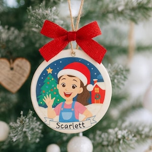 Puede incluir: Adorno navideño redondo con lazo rojo y cuerda para colgar. El adorno presenta una ilustración de dibujos animados de una niña con gorro de Papá Noel, un árbol de Navidad y el nombre "Scarlett".