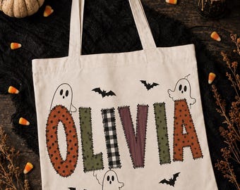 May include: A cream-colored canvas tote bag featuring the name OLIVIA in large, stitched-style letters with various patterns including orange polka dots, green, black and white buffalo plaid, and purple stripes, accented by ghost and bat illustrations.