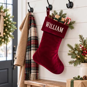 May include: A deep red velvet Christmas stocking featuring a white cuff embroidered with the name WILLIAM in capital letters. The stocking is filled with pine branches, a small gift box wrapped in brown paper, and a red and white candy cane.