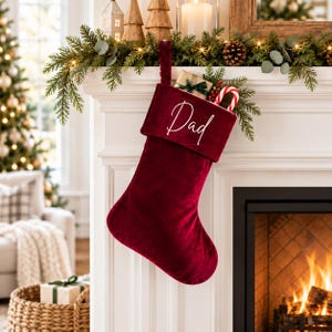 May include: A deep red velvet Christmas stocking featuring the word Dad embroidered in white script on the cuff. The stocking is filled with a small gift wrapped in brown paper and a red and white striped candy cane.