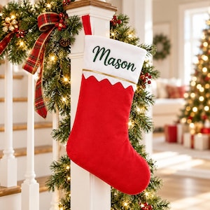 May include: A red Christmas stocking with a white cuff featuring the name Mason embroidered in dark green script. The cuff has a gold trim line and a scalloped bottom edge. The stocking is hung from a white wooden banister post.