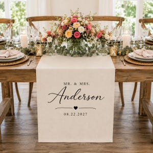 Puede incluir: Un camino de mesa de tela beige con el texto impreso en negro MR. & MRS. Anderson, un pequeño icono de corazón y la fecha 22.08.2027. El camino está colocado sobre una mesa de madera con centros de mesa florales y vajilla dispuesta.