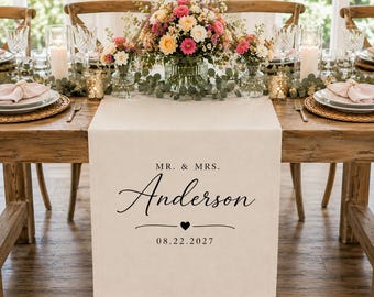 Może przedstawiać: Beżowy bieżnik z tkaniny z czarnym nadrukiem MR. & MRS. Anderson, małą ikoną serca i datą 22.08.2027. Bieżnik jest rozłożony na drewnianym stole, na którym znajdują się kwiatowe dekoracje, szklane naczynia oraz przygotowane nakrycia.