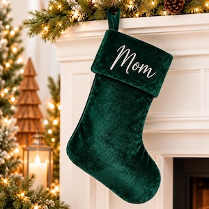 May include: A deep forest green velvet Christmas stocking featuring a folded cuff at the top. The word Mom is embroidered in white cursive script on the front of the cuff. The stocking includes a matching green velvet loop for hanging.