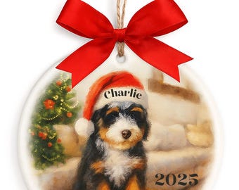 May include: A round ceramic Christmas ornament featuring a puppy wearing a Santa hat with the name "Charlie". The ornament has a red bow and the year "2025" printed on it. The background has a winter scene.