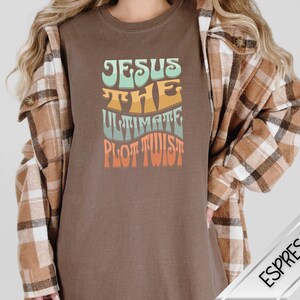 May include: Espresso-colored t-shirt with the text "JESUS THE ULTIMATE PLOT TWIST" in a retro font. The shirt is a solid color and has a crew neck. The model is wearing a plaid shirt over the t-shirt.