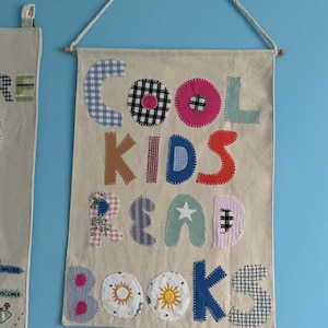 Op de afbeelding: Een rechthoekige wandhanger van beige stof met de geappliqueerde tekst COOL KIDS READ BOOKS in diverse kleurrijke patronen en texturen, waaronder vichy, bloemen en stippen, opgehangen aan een houten stok met een wit touw.