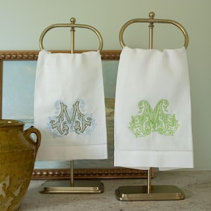 May include: Two white hand towels with embroidered monograms hanging on brass stands. One towel features a blue and grey letter M, while the other displays a green letter M. A mustard yellow ceramic pitcher sits to the left of the stands.