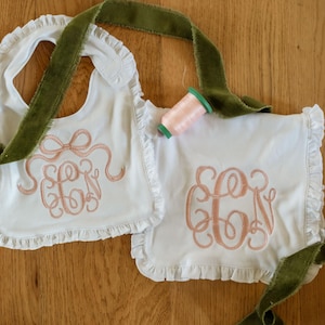 Op de afbeelding: Twee witte baby slabbetjes met geschulpte randen en geborduurde initialen in een scriptlettertype. Een roze strik ontwerp staat op één slab. Groene fluwelen linten en een spoel roze garen zijn ook aanwezig.