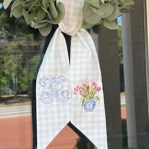 以下が含まれることがあります： 水色と白のギンガムチェックのドアハンガー。刺繍のディテールが施されています。デザインにはモノグラムとピンクのチューリップの花瓶が含まれています。ドアハンガーは緑色のユーカリのリースに結ばれています。