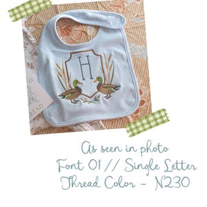 Op de afbeelding: Een lichtblauw slabbetje met een geborduurd embleem met de letter H in het midden, geflankeerd door twee wilde eenden en hoog gras. De tekst op de afbeelding luidt: As seen in photo, Font 01 // Single Letter, Thread Color - N230.