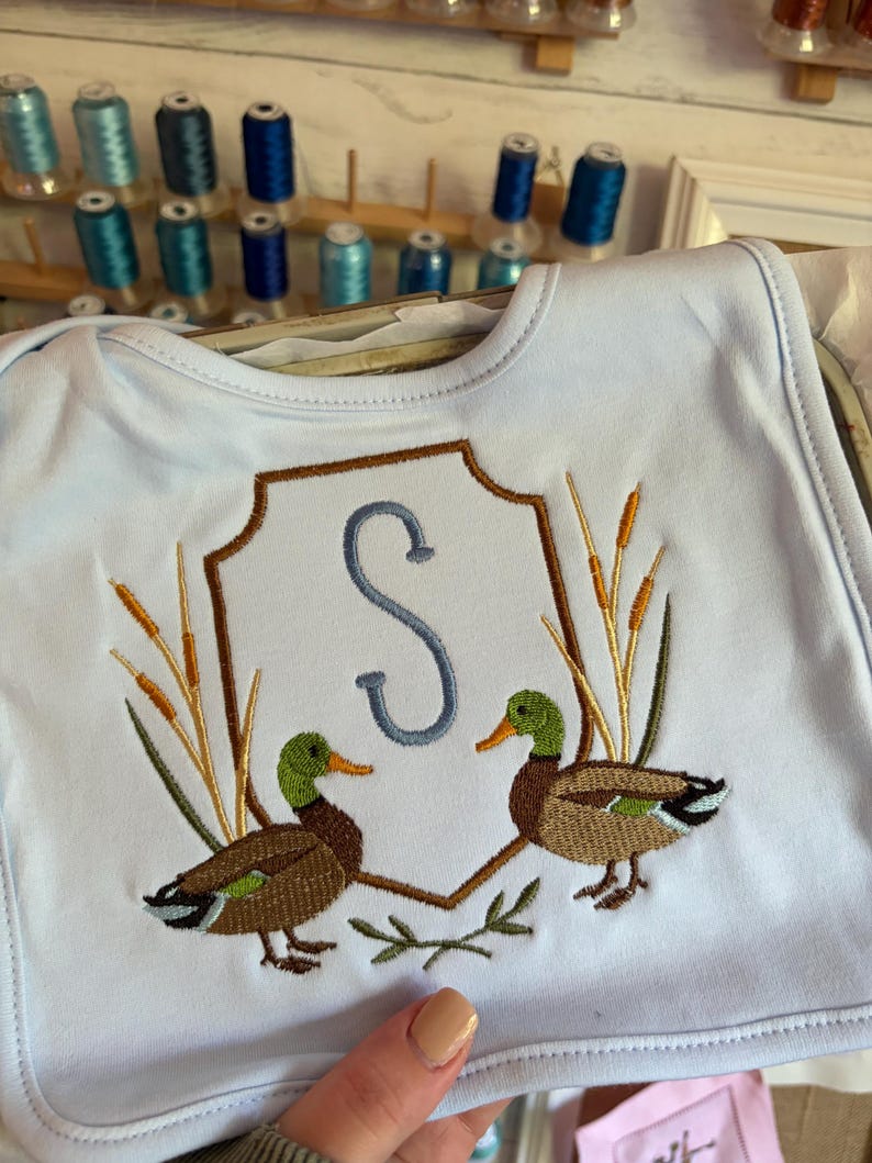 Op de afbeelding: Een lichtblauw slabbetje met een borduursel van twee wilde eenden die naar elkaar toe kijken. Tussen de eenden staat een bruine schildomtrek met een grote, lichtblauwe letter S. Hoge lisdodden flankeren het schild aan beide kanten.