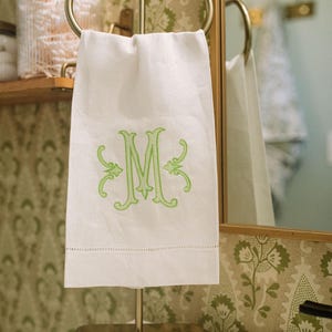 Op de afbeelding: Een witte handdoek met een limoengroene geborduurde monogramletter M in een decoratief schreeflettertype, hangend aan een goudkleurige metalen standaard. De handdoek heeft een ajourzoomdetail bij de onderrand en een gladde, schone textuur.