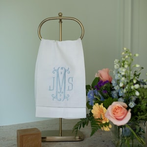 May include: A white hand towel with blue embroidered initials hangs on a gold-coloured towel rack. A bar of soap and a vase of flowers are also in the image. The towel features the initials "JMS" in a decorative font.
