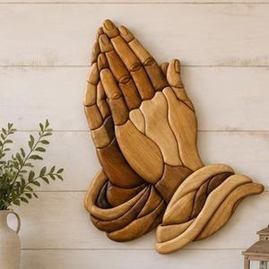 Può includere: Un'opera d'arte da parete in legno che raffigura due mani giunte in posizione di preghiera. La scultura è composta da molteplici segmenti di legno a incastro in varie tonalità di marrone naturale, creando un effetto tridimensionale a strati.