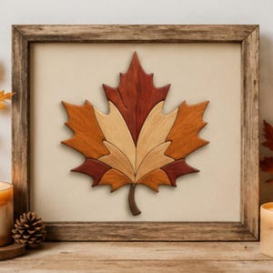 May include: A framed wall art piece featuring a central maple leaf design composed of segmented wooden pieces in varying shades of deep red, orange, and light tan, set against a neutral background within a rustic brown wooden frame.