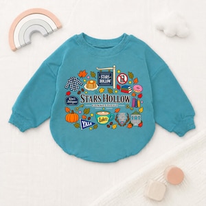 Könnte beinhalten: Ein blaugrünes Langarm-Sweatshirt mit einem Grafikdruck mit dem Text Stars Hollow Connecticut, Luke's, Kim's Antiques, Yale und I Got Hit By A Deer, umgeben von Illustrationen von Herbstblättern, einem Kürbis, Kaffee und Büchern.