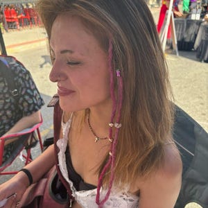 May include: A hair accessory consisting of two long, magenta-colored yarn strands braided into brown hair. The strands are adorned with small, metallic gold-toned beads and a single purple cylindrical bead, hanging down alongside the face.