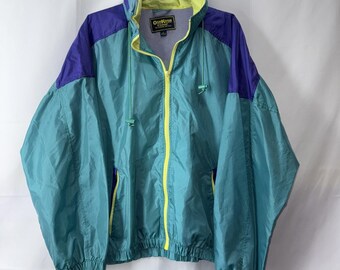 Könnte beinhalten: Eine Vintage-Windjacke in Türkis, Lila und Gelb. Die Jacke hat einen durchgehenden Reißverschluss, eine Kapuze und Taschen. Der Markenname "OshKosh" ist auf dem Etikett zu sehen. Die Jacke hängt an einem Metallbügel.