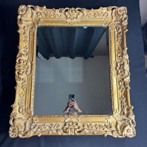 May include: A rectangular wall mirror featuring a thick, ornate gold-colored frame with intricate relief carvings of floral and scroll motifs. The reflective glass surface is clear and smooth, set within the detailed, textured metallic border.