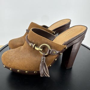 May include: A pair of brown suede mules with dark brown leather trim, featuring a brass-toned metal ring detail, a hanging leather tassel, and a high block heel. The base of the shoe is accented with small brass-toned studs along the perimeter.