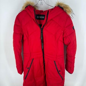 May include: A bright red Guess winter coat with a hood and faux fur trim. The coat has a black zipper, black trim on the pockets, and the Guess logo on the chest. The coat is hanging on a metal hanger.