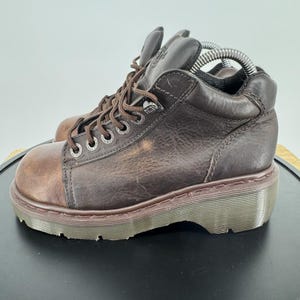 Può includere: Stivaletti in pelle marrone con punta arrotondata e suola spessa e scanalata. Gli stivali hanno lacci marroni e occhielli metallici. La punta è di una tonalità di marrone più chiara.