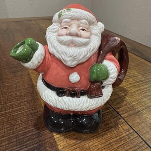 Könnte beinhalten: Eine Keramikfigur des Weihnachtsmanns mit rotem Mantel, schwarzen Stiefeln und weißem Bart. Die Figur hält einen braunen Sack und einen grünen Fäustling. Der Weihnachtsmann hat einen fröhlichen Ausdruck und ist mit Stechpalmen verziert.